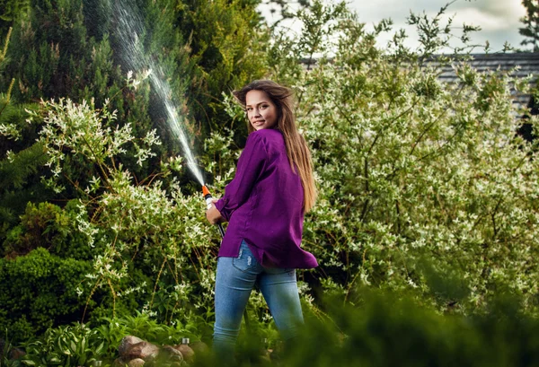 Beautiful joyful young girl in violet casual shirt poses in a summer garden with water hose. — Zdjęcie stockowe