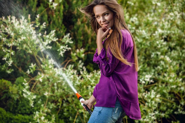 Beautiful joyful young girl in violet casual shirt poses in a summer garden with water hose. — Zdjęcie stockowe