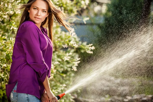Beautiful joyful young girl in violet casual shirt poses in a summer garden with water hose. — Stock fotografie
