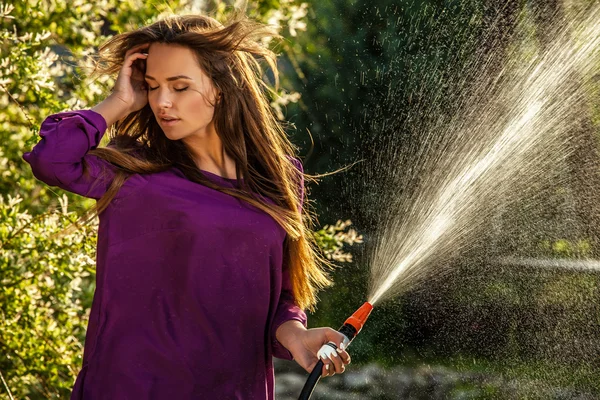 Schöne fröhliche junge Mädchen in violettem lässigem Hemd posiert in einem Sommergarten mit Wasserschlauch. — Stockfoto