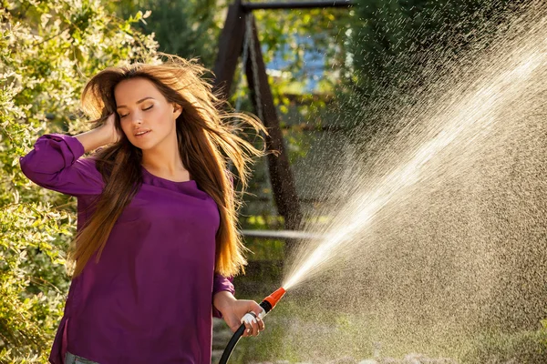 Beautiful joyful young girl in violet casual shirt poses in a summer garden with water hose. — Stock Photo, Image