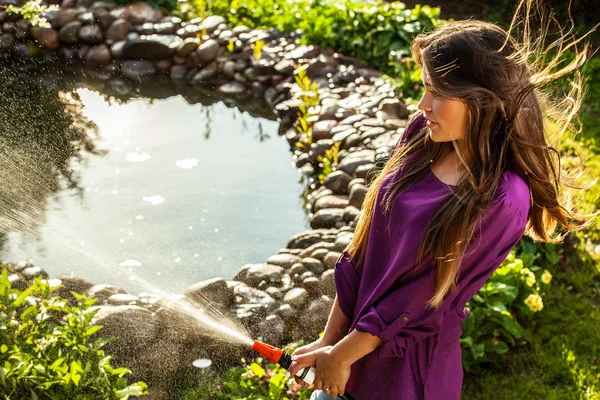 Schöne fröhliche junge Mädchen in violettem lässigem Hemd posiert in einem Sommergarten mit Wasserschlauch. — Stockfoto