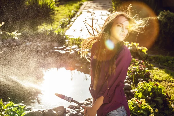 Beautiful joyful young girl in violet casual shirt poses in a summer garden with water hose. — Stock Photo, Image