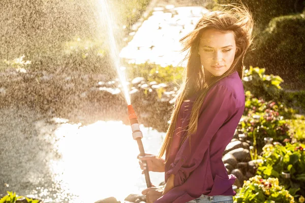 Schöne fröhliche junge Mädchen in violettem lässigem Hemd posiert in einem Sommergarten mit Wasserschlauch. — Stockfoto