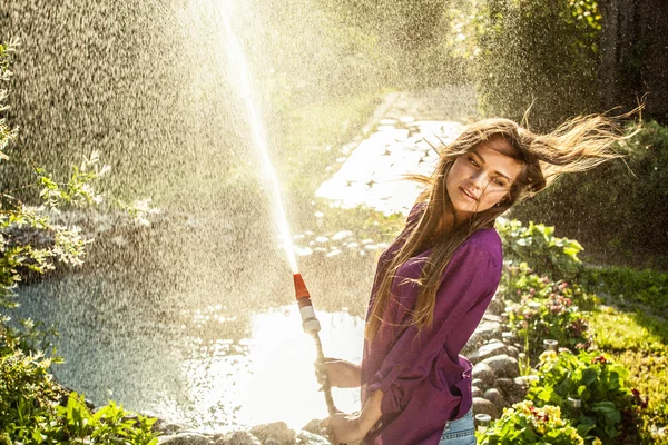 Schöne fröhliche junge Mädchen in violettem lässigem Hemd posiert in einem Sommergarten mit Wasserschlauch. — Stockfoto