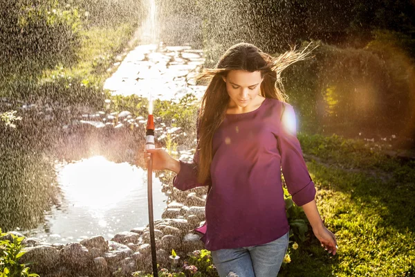 Beautiful joyful young girl in violet casual shirt poses in a summer garden with water hose. — Stock Photo, Image