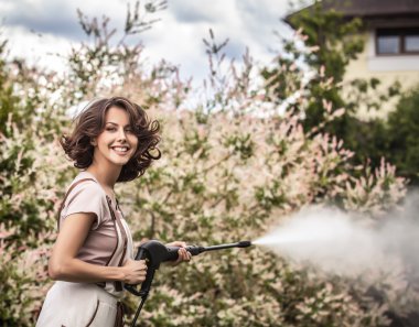 Outdoors portrait of beautiful young woman in overalls which posing with water-cannon in summer garden.