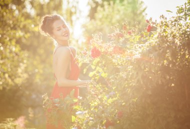 Outdoors portrait of beautiful young brunette girl in luxury red dress posing in summer garden.