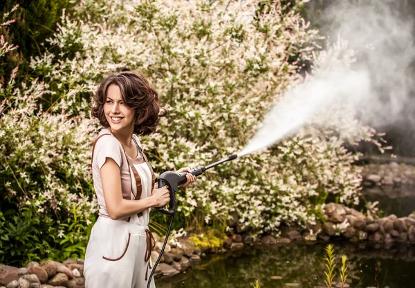 Outdoors portrait of beautiful young woman in overalls which posing with water-cannon in summer garden. — Stok fotoğraf