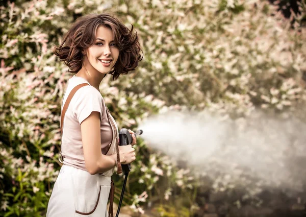 Outdoors portrait of beautiful young woman in overalls which posing with water-cannon in summer garden. — Zdjęcie stockowe