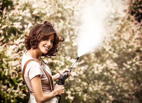 Outdoors portrait of beautiful young woman in overalls which posing with water-cannon in summer garden. — ストック写真