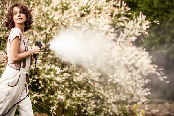 Outdoors portrait of beautiful young woman in overalls which posing with water-cannon in summer garden. — Stok fotoğraf