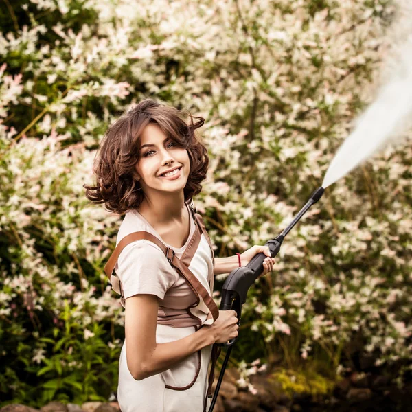 Outdoors portrait of beautifu young woman in overalls which posing with water-cannon in summer garden. — Stock Photo, Image