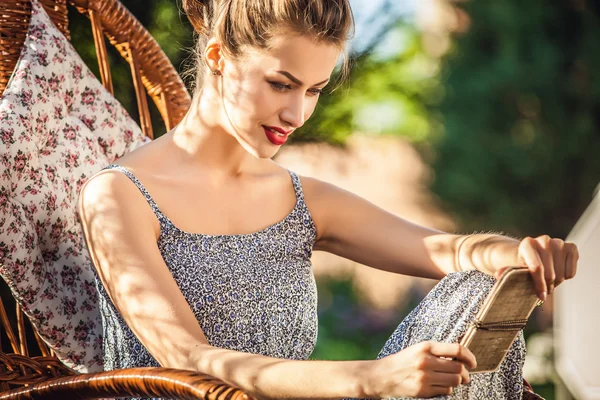 Outdoors portrait of beautiful young woman which sitting in rattan rocking-chair in evening summer garden. — Zdjęcie stockowe