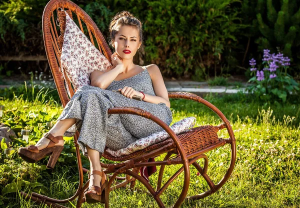 Outdoors portrait of beautiful young woman which sitting in rattan rocking-chair in evening summer garden. — Stock Photo, Image