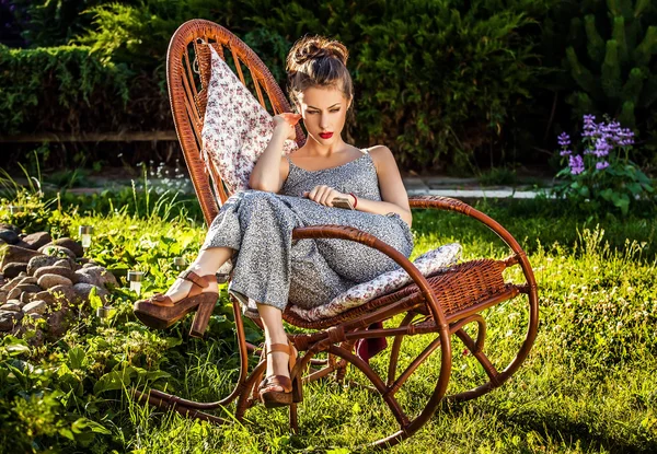 Outdoors portrait of beautiful young woman which sitting in rattan rocking-chair in evening summer garden. — Stock Photo, Image