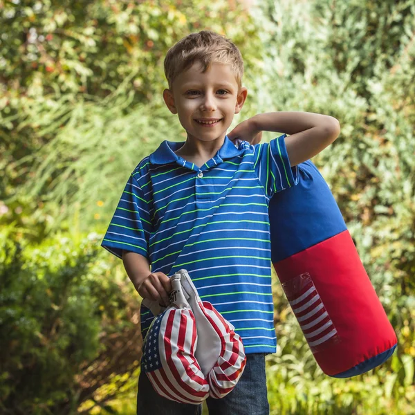 Outdoor portrait of positive little boy in sunny summer garden with children's punching bag and gloves. — Stockfoto
