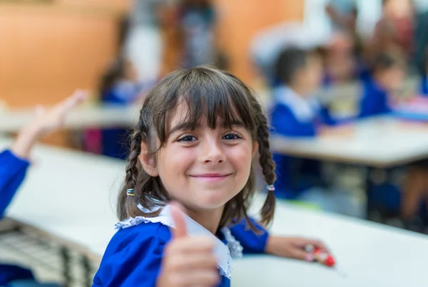 Elementary schoolgirl with thumb up in her classroom — Stock Photo, Image
