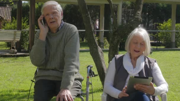Idős férfi és nő mobiltelefonnal és táblagéppel a kórház kertjében. Lassú mozgás. — Stock videók