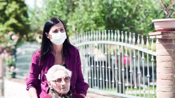 Vue de face d'une jeune femme portant un masque aidant sa mère handicapée en fauteuil roulant dans le parc de la ville — Video