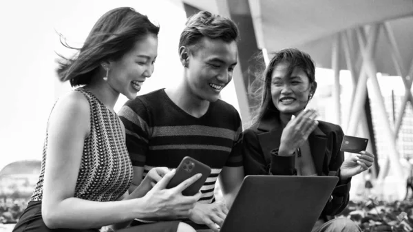 Three asian young friends seated on a bench making shoppin on the web with the laptop and credit card.