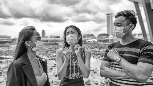 Three asian young friends standing outdoor in the city talking and wearing face masks in covid coronavirus pandemic.