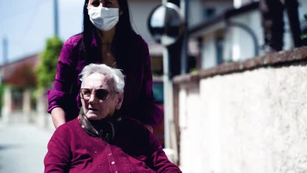 Vooraanzicht van een jonge vrouw dragen masker assisteren haar gehandicapte moeder op rolstoel in het stadspark, slow motion — Stockvideo