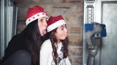 Noel 'de evde Noel şapkası takan bir kadın ve kızı kameranın önünde surat asıyorlar. Ebeveynlerle video çağrısı