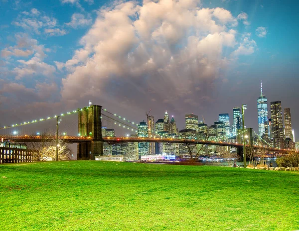 ニューヨーク市の前景の背景と緑の牧草地にマンハッタンのスカイラインと日没時のブルックリン橋 — ストック写真