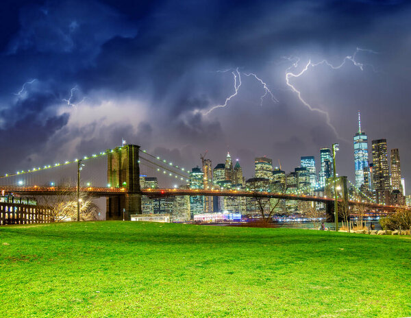 The Brooklyn Bridge during a storm with Manhattan skyline on background and green meadow in foreground, New York City.