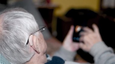 80 'li yaşlarda emekli bir adam kanepeye yaslanmış ve haber için akıllı telefonu kullanıyor.