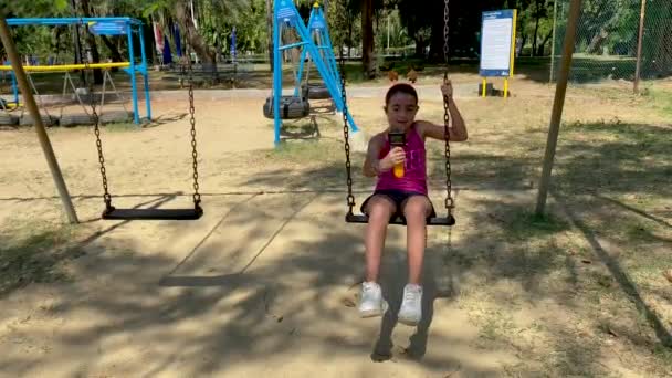 Jong meisje genieten swing in een mooie stad park, hlding compact camera — Stockvideo