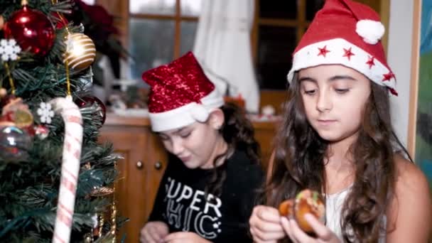 Two young girls at home decorating Christmas Tree wearing Santa Hat — Stock Video