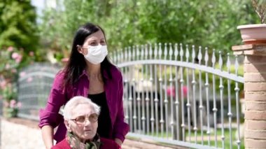 Engelli annesine parkta tekerlekli sandalyede yardım eden maskeli genç bir kadının ön görüntüsü, yavaş çekim.