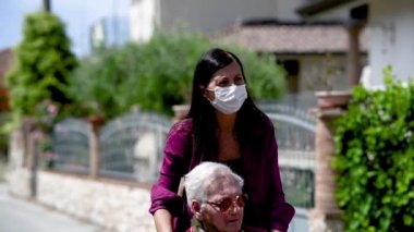 Engelli annesine parkta tekerlekli sandalyede yardım eden maskeli genç bir kadının ön görüntüsü, yavaş çekim.