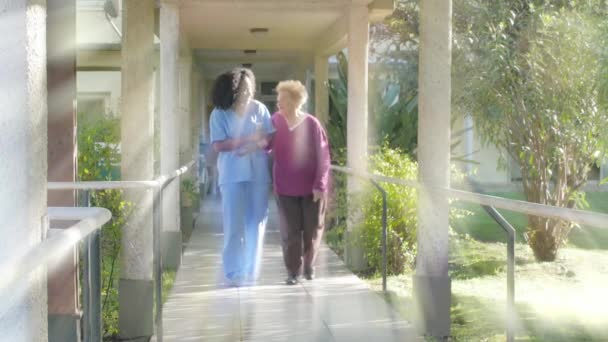 African doctor helps elderly caucasian woman walking along the rehab clinic garden, sun light rays coming from the sky — Stock Video