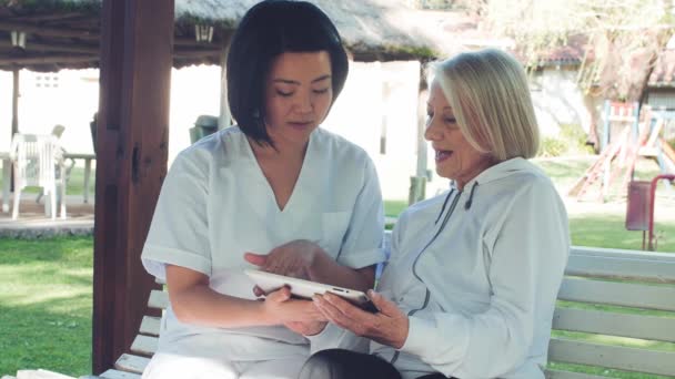 Médico asiático explicando la prueba médica en la tableta a una mujer anciana, ambos sentados en un banco — Vídeos de Stock