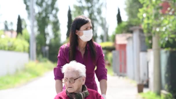 Vooraanzicht van een jonge vrouw dragen masker assisteren haar gehandicapte moeder op rolstoel in het stadspark — Stockvideo