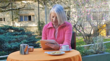 Kafkasyalı emekli kadın teknolojik aletler kullanıyor kahvaltıda bahçede dinleniyor, video görüşmesi yapıyor. Emeklilik, teknoloji ve rahatlama kavramı.