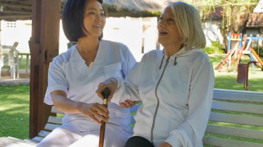 Asyalı doktor, yaşlı ve emekli bir kadınla konuşuyor. İkisi de bahçe bankında oturuyor. Mutluluk ve emeklilik kavramı.