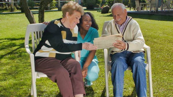 年轻的非洲女护士和退休的老夫妇一起在室外微笑 在医院花园用平板电脑与他们交谈 — 图库照片