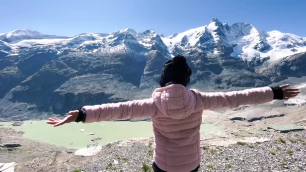 Πλάτη άποψη ενός νεαρού κοριτσιού αγκαλιάζει καθαρό αέρα με βουνά που καλύπτονται από χιόνι κατά τη θερινή περίοδο, Grossglockner Εθνικό Πάρκο, Αυστρία — Αρχείο Βίντεο