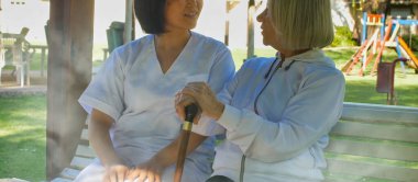 Asyalı doktor, yaşlı ve emekli bir kadınla konuşuyor. İkisi de bahçe bankında oturuyor. Mutluluk ve emeklilik kavramı.