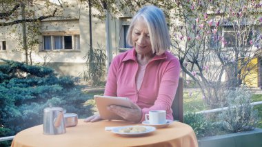 Kafkasyalı emekli kadın teknolojik aletler kullanıyor kahvaltıda bahçede dinleniyor, video görüşmesi yapıyor. Emeklilik, teknoloji ve rahatlama kavramı.