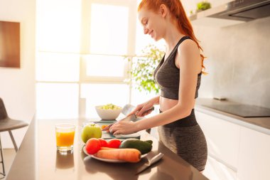 Atletik genç kızıl saçlı kadın ev mutfağında sebze santrifüjlü ve salata hazırlıyor.