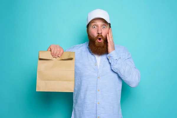 Deliveryman con espressione stupita pronto a consegnare borsa con il cibo. sfondo ciano. — Foto Stock