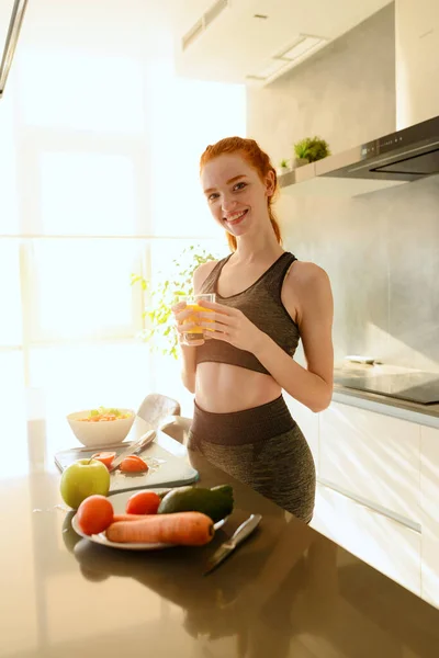 Athletic woman with gym clothes drinks orange fruit in the kitchen