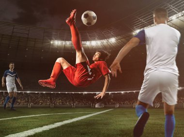 Kırmızı üniformalı bir futbolcu stadyumda topa akrobatik bir tekmeyle vuruyor.