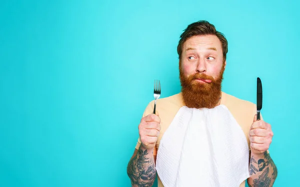 Człowiek z tatuażami jest gotowy do jedzenia ze sztućcami w ręku. tło cyjanu — Zdjęcie stockowe