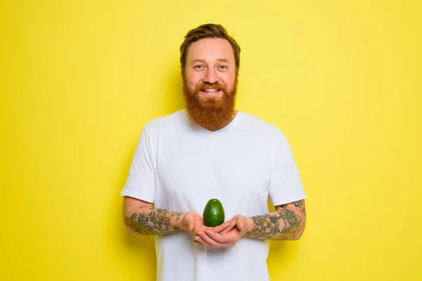 Uomo felice con la barba e tatuaggi tiene un avocado — Foto Stock